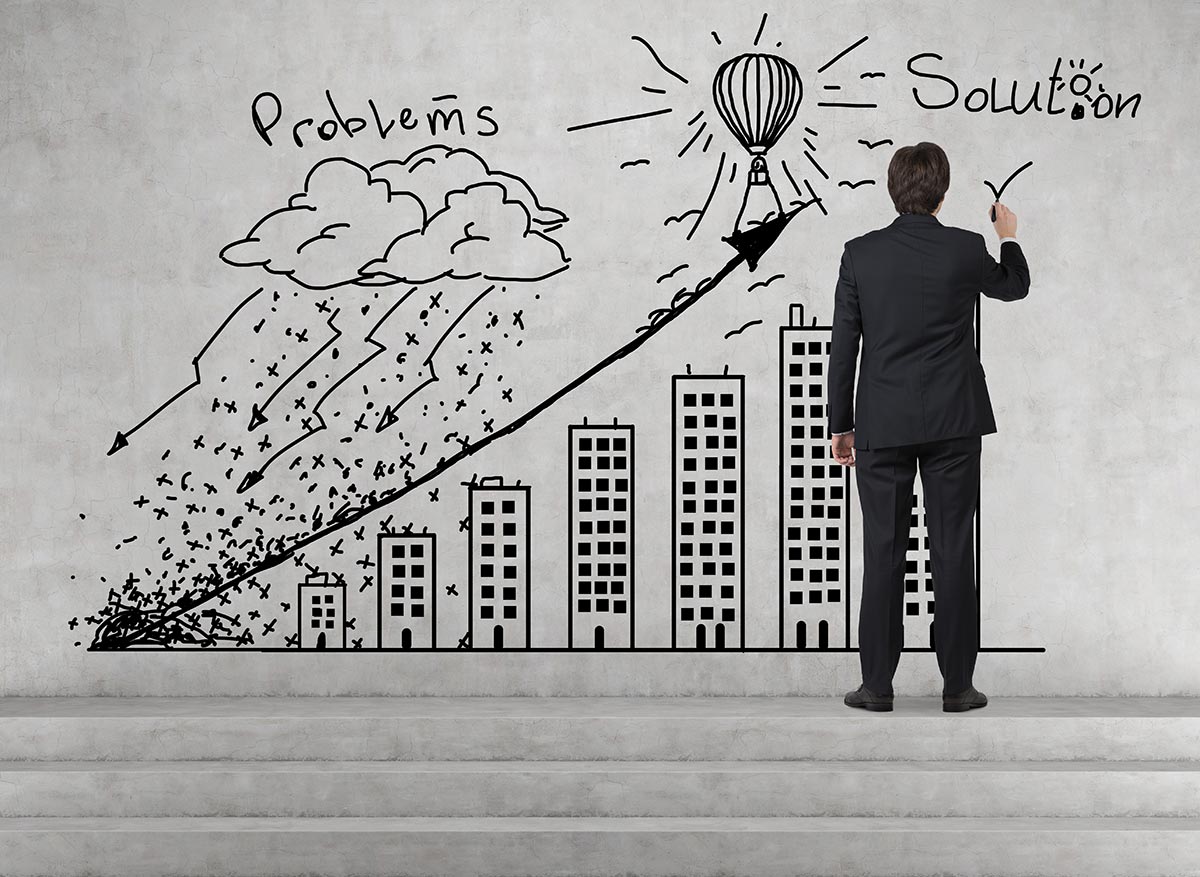 A man drawing a picture on the wall of sky scrapers that get taller from left to right with a line above the skyscrapers with an arrow at the end. At the top left it has the word Problems with rain clouds below the word, and at the top right it has the word solution with the sun as the dot to the letter i. In between the words Problems and Solution is a drawing of a hot air balloon